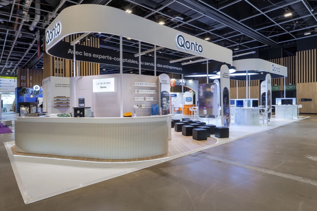 Stand Qonto au Congrès de l'Ordre des Experts-Comptables, bar arrondi « L3 B1STRO », poufs noirs et postes de démonstration sous un bandeau blanc lumineux, conçu et réalisé par The Good Experience
