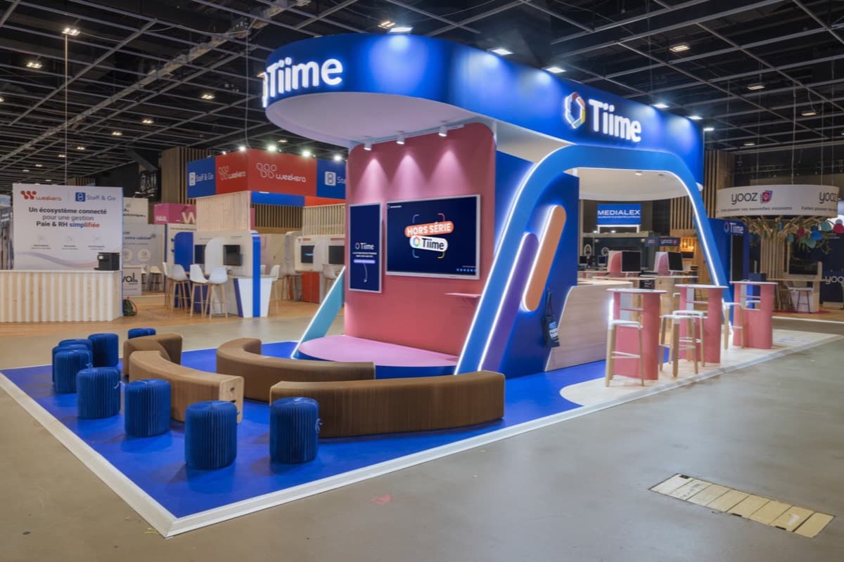 Stand Tiime au Congrès de l'Ordre des Experts-Comptables, scène vidéo rose sous un bandeau bleu lumineux, banquettes en carton et tables hautes, conçu et réalisé par The Good Experience